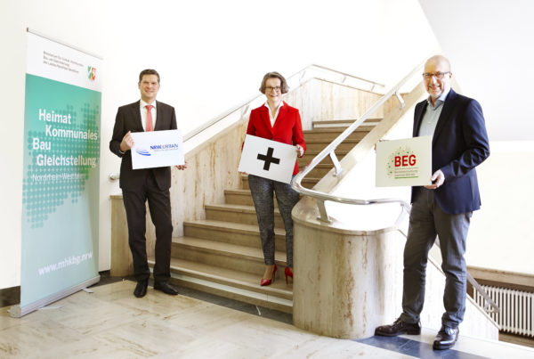 Herr Brockemeyer, Ministerin Frau Scharrenbach, Herr Kloidt halten Schilder die das zusammenwachsen von BEG und NRWA Urban symbolisieren