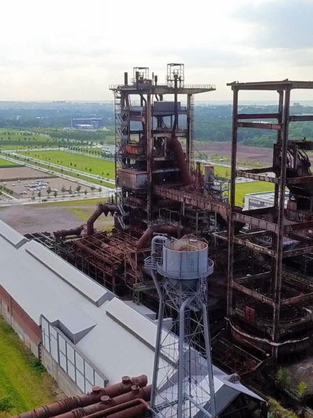 Luftaufnahme mit einer alten Industrieanlage in der Bildmitte. An den Seiten Wiesen, Bäume, Straßen, Parkplätze mit parkenden Autos und neuere Gebäude. Im Hintergrund weitere Grünflächen und ein Hochhaus.