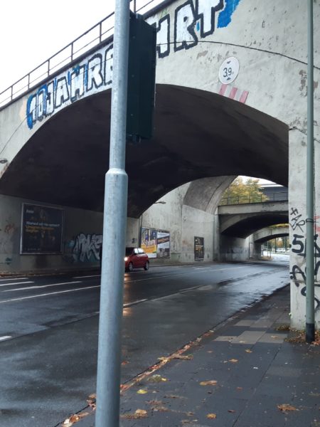 Eine Straße, über die eine Überführung verläuft. Auf der Straße fahren ein rotes und silberfarbenes Auto. Auf der Überführung befinden sich Graffitis.