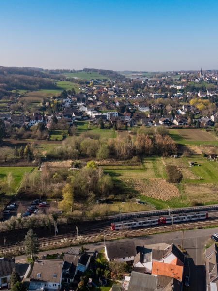 Luftaufnahme einer Kleinstadt im Grünen mit Wiesen, Bäumen, Wohnhäusern und Schienenverlauf im vorderen Bildteil. Darüber blauer Himmel.