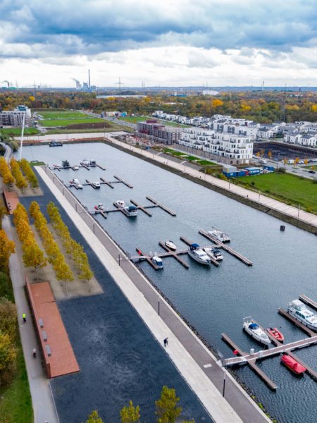 Am linken Bildrand verläuft ein Fluss, der mittig in einen kleinen Hafen mit Bootsanlegern und Booten mündet. Im rechten und hinteren Bildteil Wohnhäuser, Straßen, Autos und Wiesen.