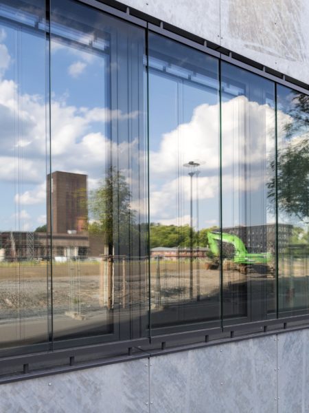 In einer Glasfront spiegeln sich mehrere Industriegebäude, ein grüner Bagger, ein Baum und die Wolken am Himmel.