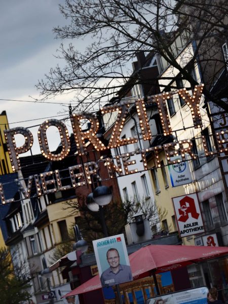 Eine Straßenzeile mit Wohnhäusern, Geschäften und einer Kirche am Ende. Quer darüber gespannte Leuchtschrift "Porzity".