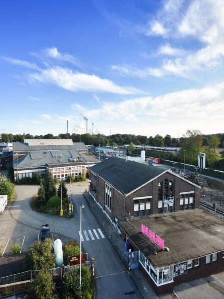 Links eine Straßengabelung, mehrere Gebäude und Bäume. Rechts ein gewerbliches Gebäude, nach rechts von Schienen gesäumt.