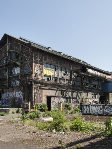 Leerstehendes Fabrikgebäude, mit Graffitis besprüht und von wildem Pflanzenwuchs umgeben.