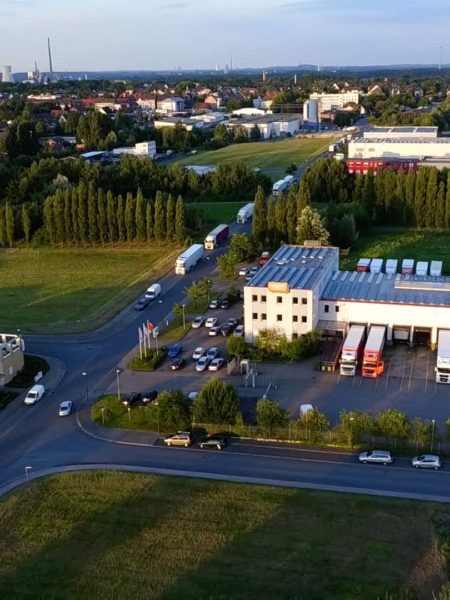 Mittig im Bild eine Straßenkreuzung, links und rechts davon gewerbliche Gebäude und parkende LKWs. Im Hintergrund Baumreihen und dahinter weitere Gewerbegebäude und Wohnhäuser.