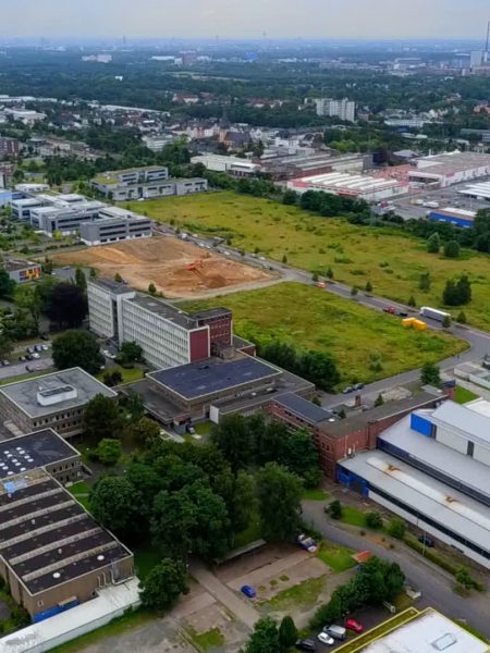 In der Mitte befinden sich ein Industriegebiet, Parkplätze, ein Baugrundstück und eine Wiese. Am linken, rechten und oberen Bildrand Wohnhäuser.
