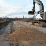 Das Foto zeigt eine Baustelle, im Vordergrund ist ein Erdhaufen zu sehen, am rechten Bildrand ein großer Bagger, im Hintergrund sieht man eine ebene, erdige Fläche.