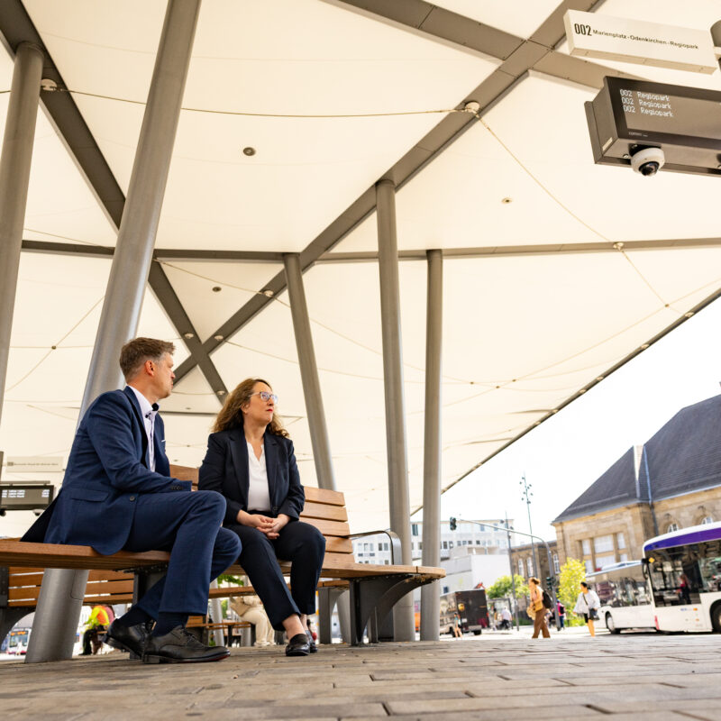 Henk Brockmeyer von NRW.URBAN und Claudia Schwan-Schmitz sitzen auf einer Bank vor dem Bahnhof Mönchengladbach